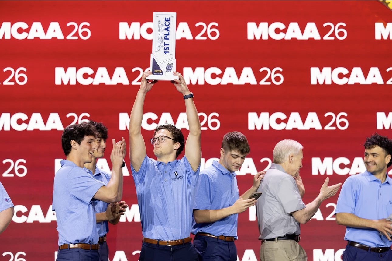 Several people wearing matching light blue polo shirts stand on a stage in front of a red backdrop displaying “MCAA 26.” One person at the center holds a first-place trophy overhead while others nearby applaud.