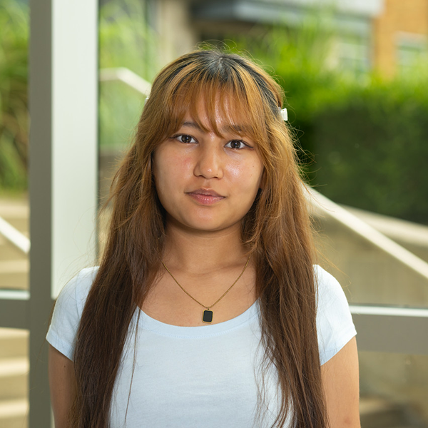 Female student with dark brown hair standing indoors near large windows in a campus building. The person is wearing a light blue shirt and a necklace, with the building exterior and greenery visible through the windows behind them.