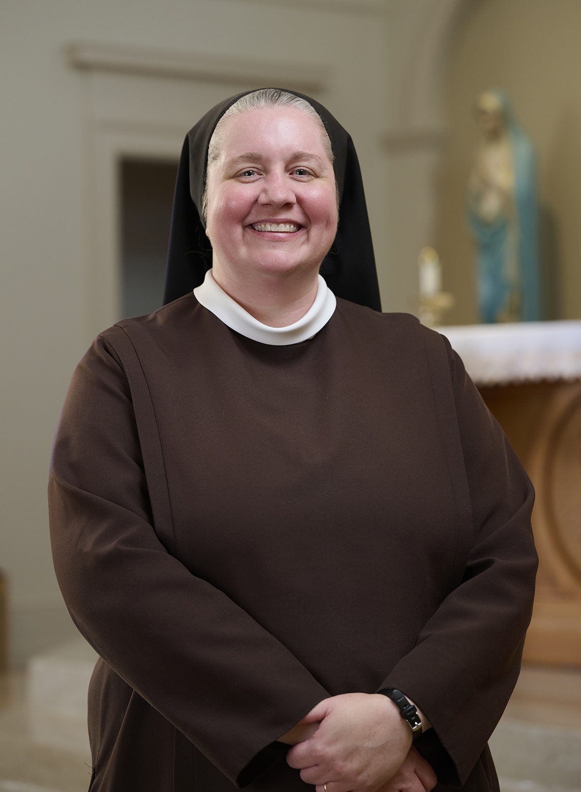 Sister Maria Gemma Salyer, OSF, smiles for a portrait while wearing a brown religious habit and black veil. She stands with her hands clasped in front of her inside a chapel.