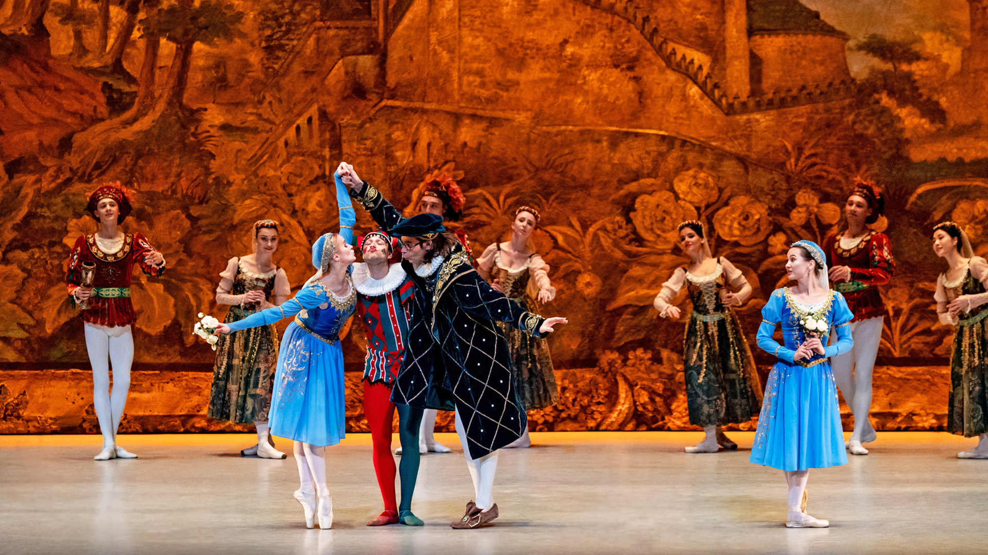 A group of ballet performers on stage in Renaissance-style costumes, featuring a central jester in a red and blue costume flanked by dancers in blue dresses and a nobleman in a dark coat, against a painted golden-brown backdrop.