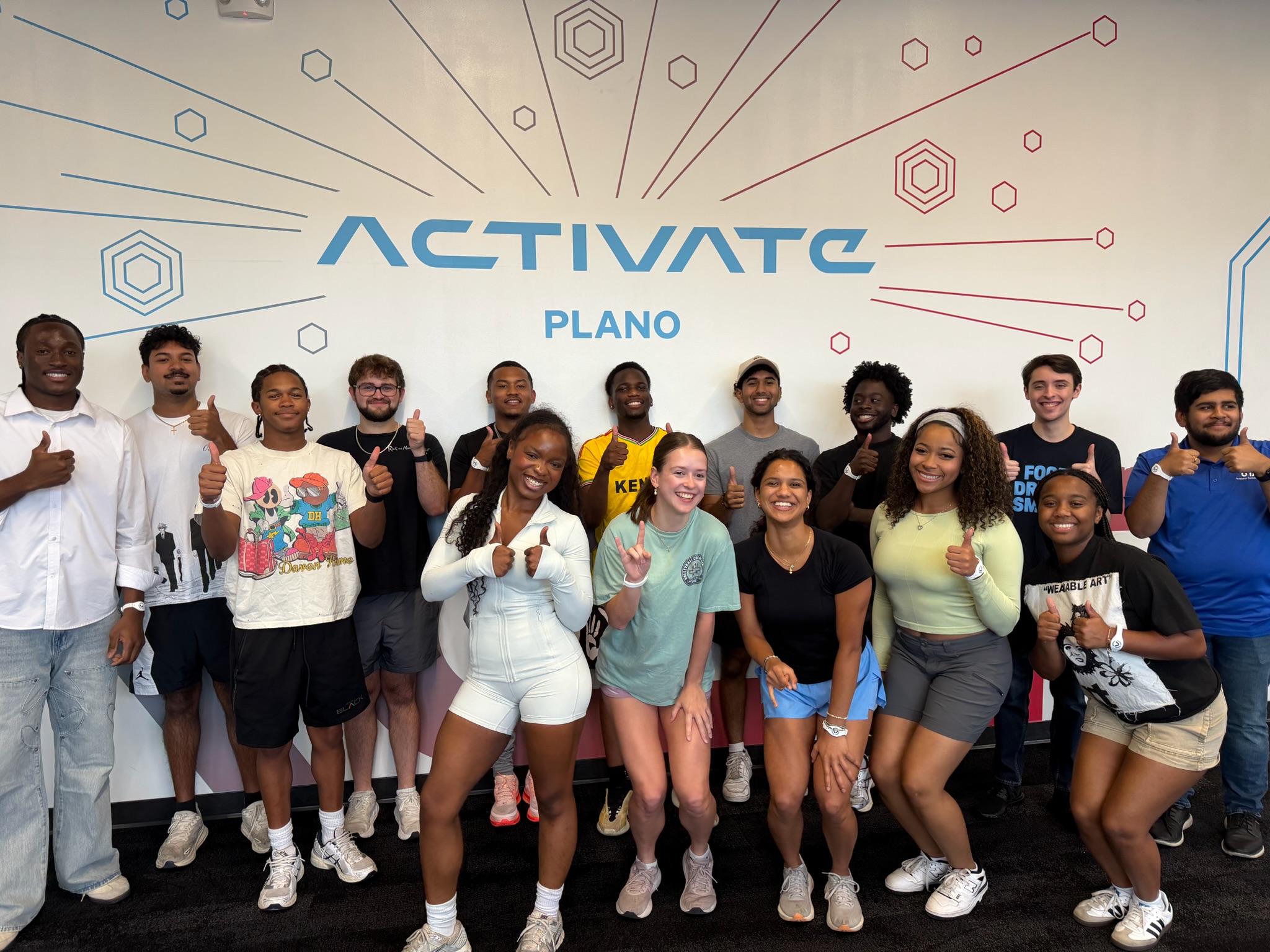 A group of students poses together, many giving a thumbs-up, in front of a wall displaying the words “Activate Plano.”