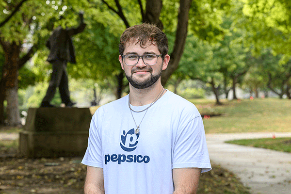 Portrait of a person standing outdoors in a park-like setting with trees and a walking path in the background. The person is wearing glasses, layered necklaces, and a light-colored T-shirt displaying the PepsiCo logo and text. A statue on a pedestal is visible in the background to the left, partially obscured by trees. The image is well lit with green foliage filling much of the background.