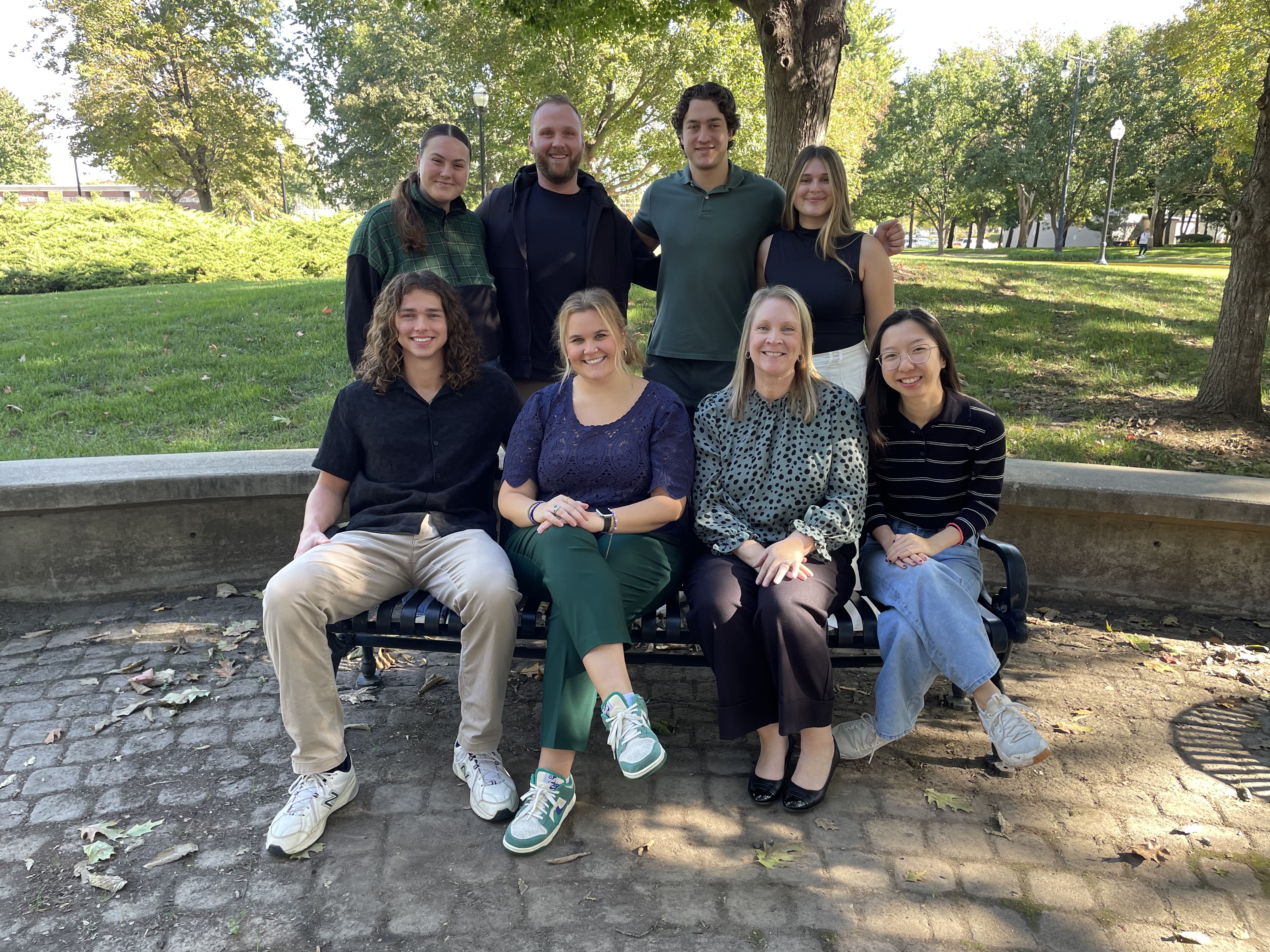 Group photo of eight students and faculty members posing outdoors on a sunny day. Four individuals are seated on a park bench over cobblestone, and four are standing behind them, with trees, a grassy lawn, and park lamps visible in the background.