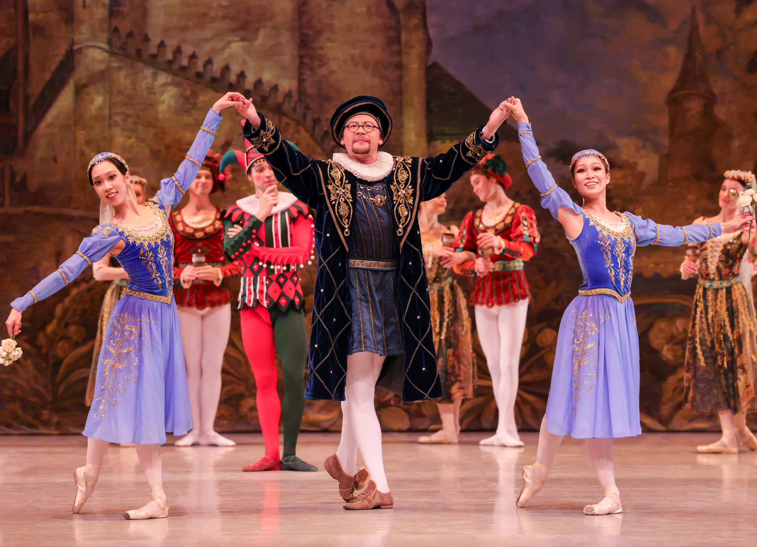 A central male performer in a dark blue velvet coat and beret holds hands with two female ballet dancers in matching blue and gold dresses, with an ensemble cast in historical costumes standing in the background against a painted castle backdrop.
