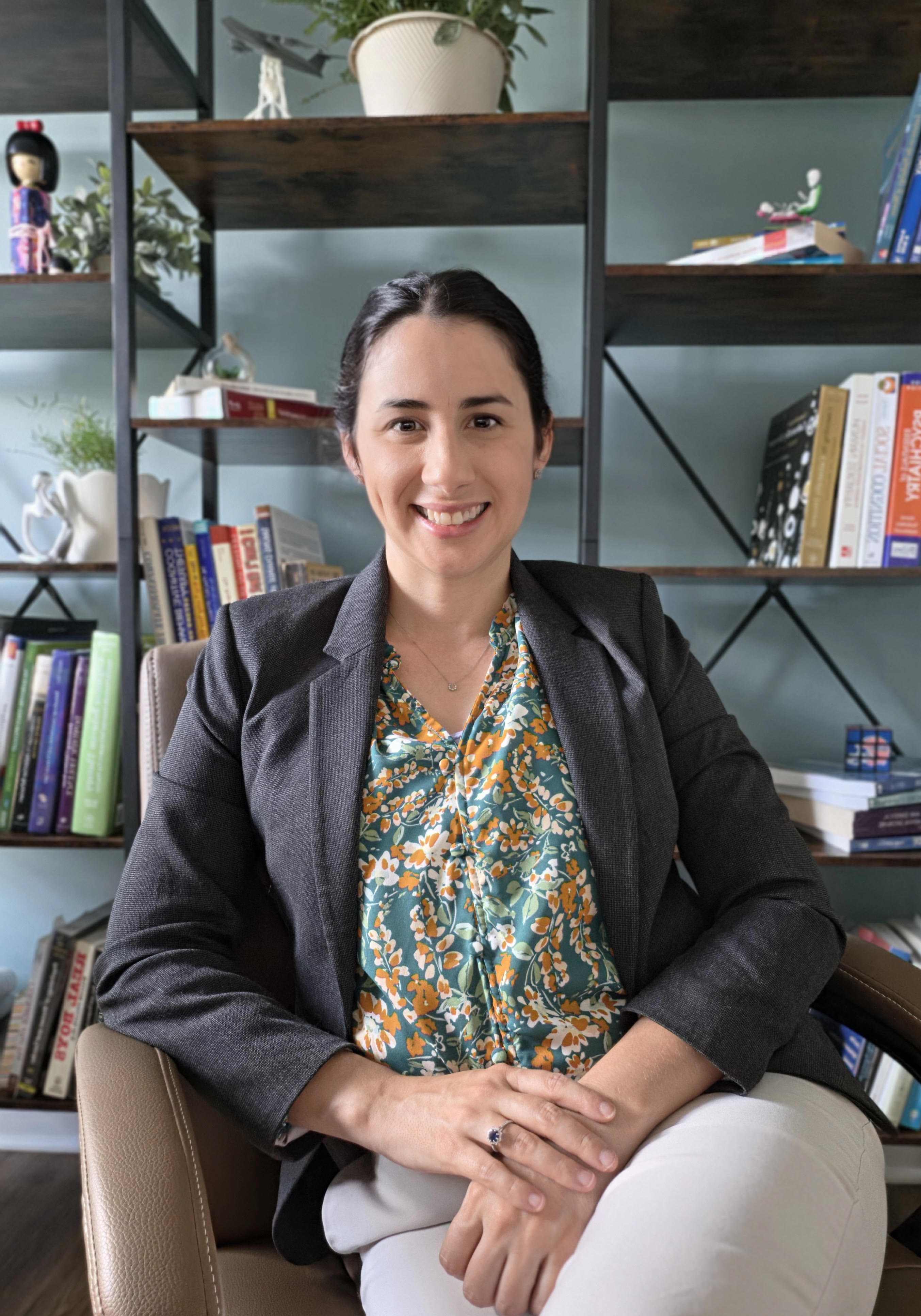 A professional headshot of a woman sitting in a chair in front of a bookshelf, wearing a dark gray blazer over a green and orange floral blouse, with her hands clasped in her lap and smiling at the camera.