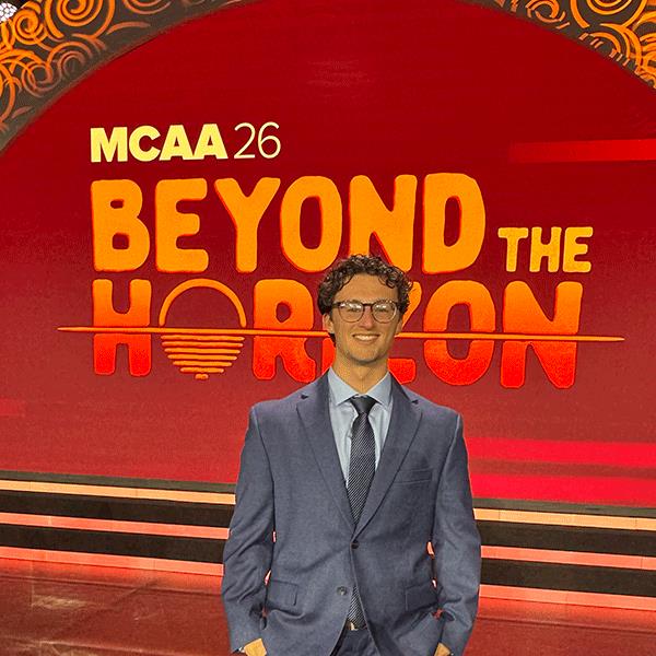 A person wearing a blue suit, white shirt, and dark tie stands in front of a large red conference stage backdrop. The backdrop displays the text “MCAA 26” and “BEYOND THE HORIZON” in large orange and yellow letters. Decorative patterned elements appear along the top edge of the stage, and illuminated horizontal stage lights run behind the person. The photo appears to be taken at a professional conference or event venue.