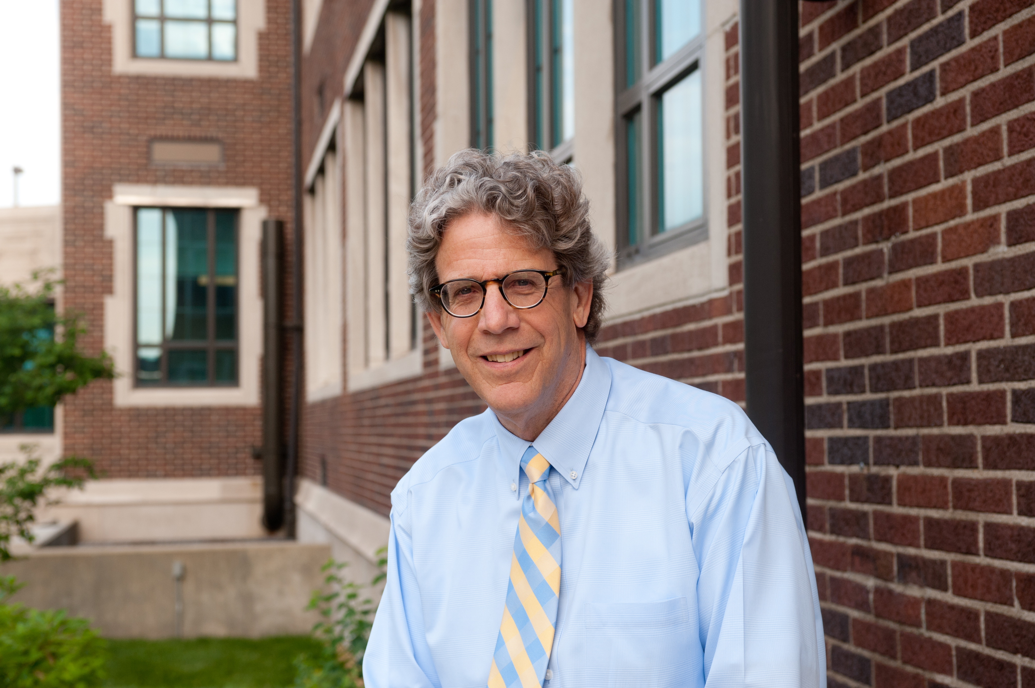 John Beacon, smiling in a light blue shirt and blue-and-gold striped tie, standing outside a brick campus building