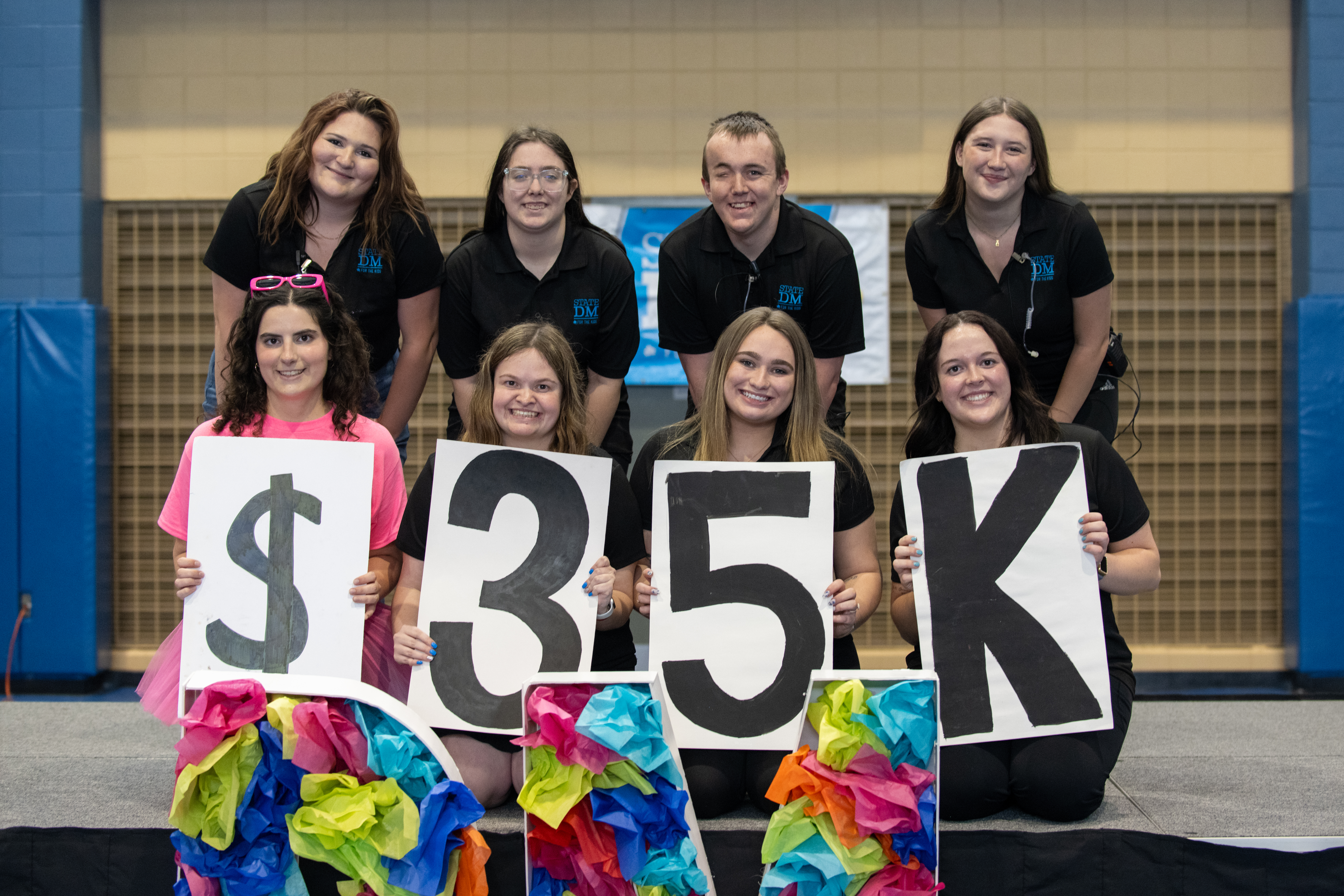 Eight young adults wearing black or pink shirts holding signs showing $35K with colorful tissue paper decorations on stage in a gymnasium.