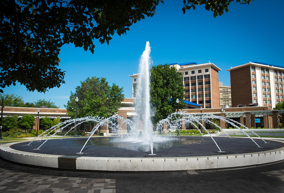 Large circular water fountain with multiple streams in front of Indiana State University buildings under clear blue sky.