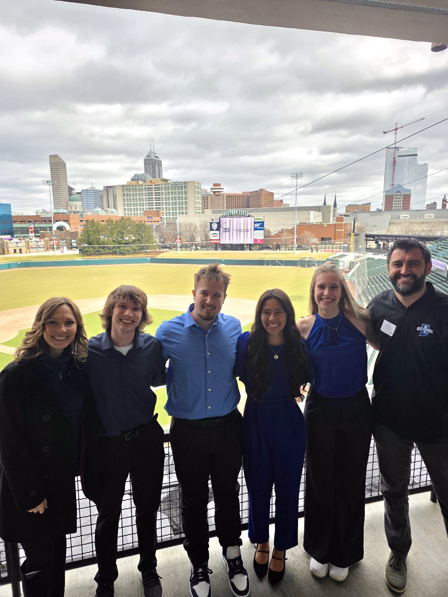 Six young adults and one older adult stand smiling in a row at a baseball stadium with a city skyline in the background under cloudy sky.