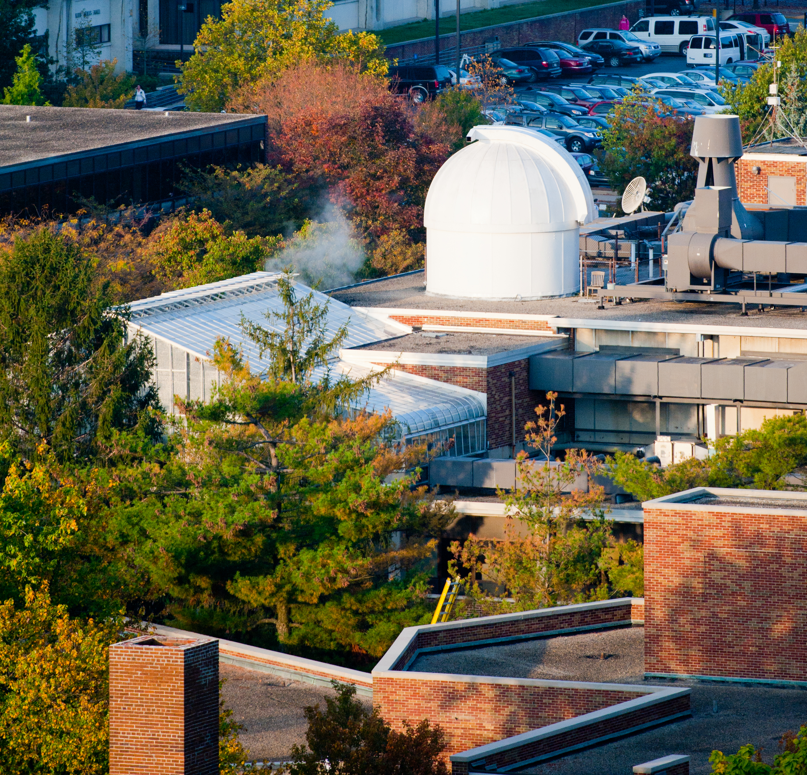 White observatory dome on a brick building rooftop surrounded by trees showing autumn colors with parked cars in the background.