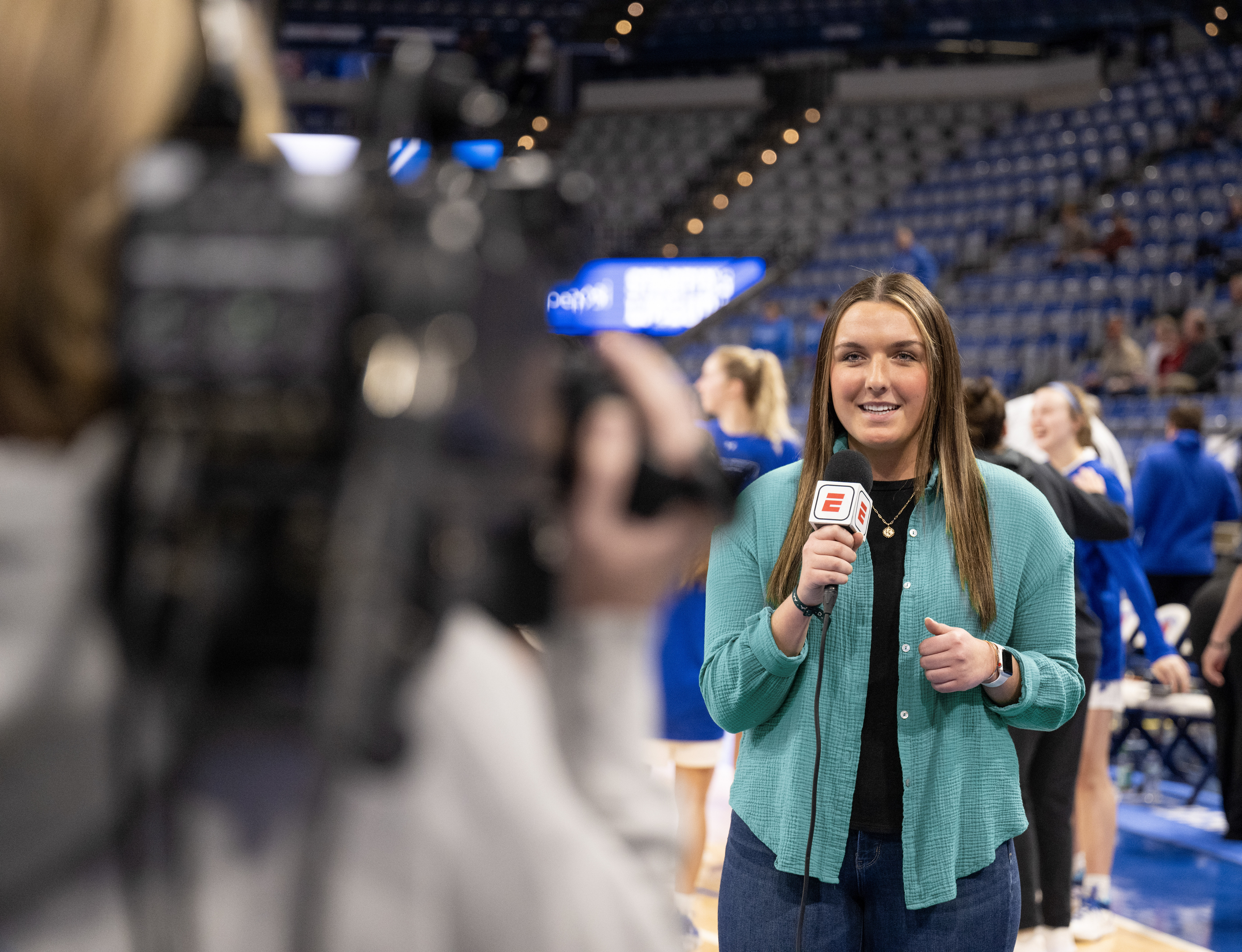 Young woman wearing a teal shirt holding an ESPN microphone, reporting in a sports arena with basketball players and spectators in the background.