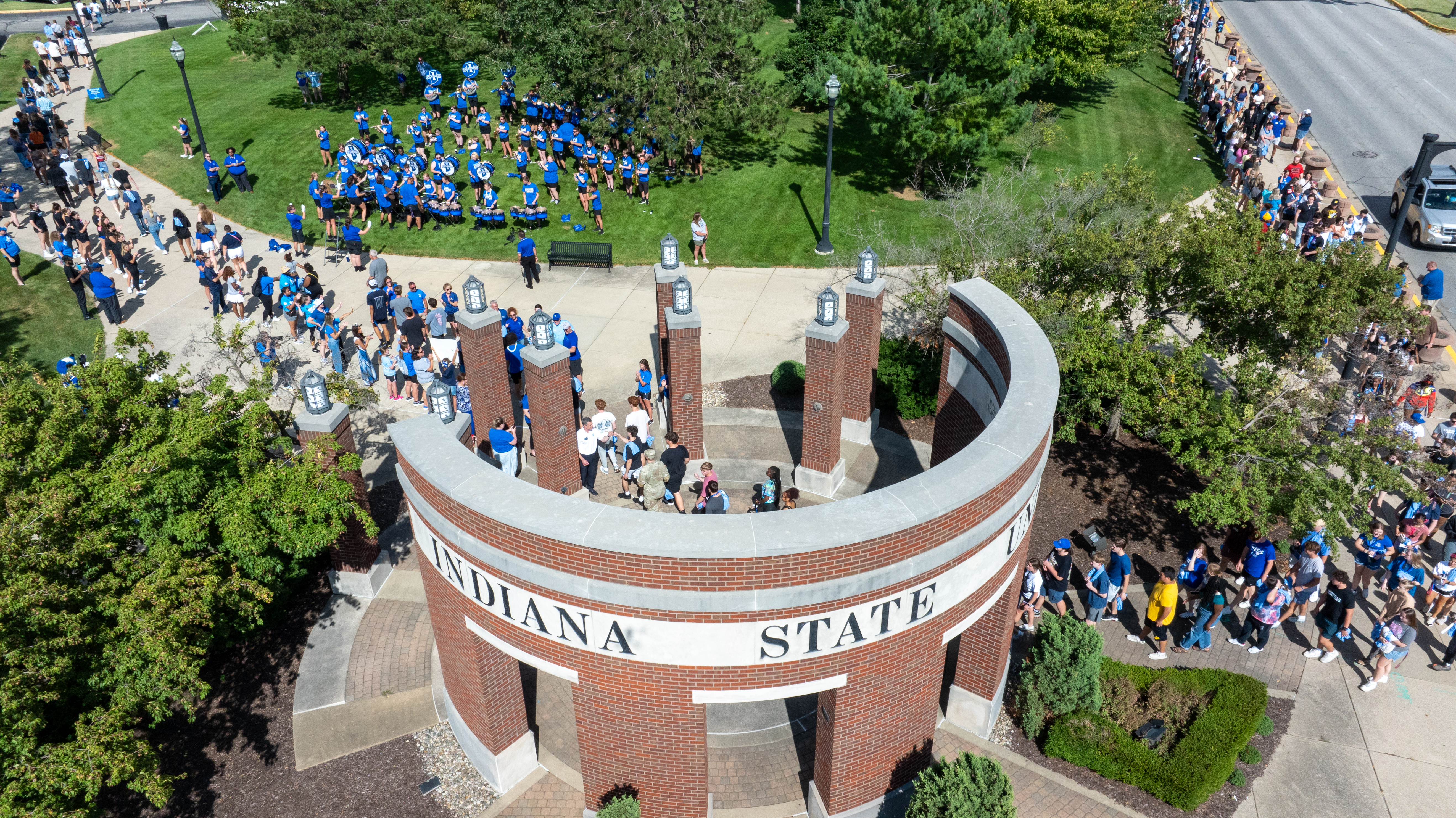 Crowd of people and marching band around the Indiana State University campus entrance sign in a sunny outdoor setting.