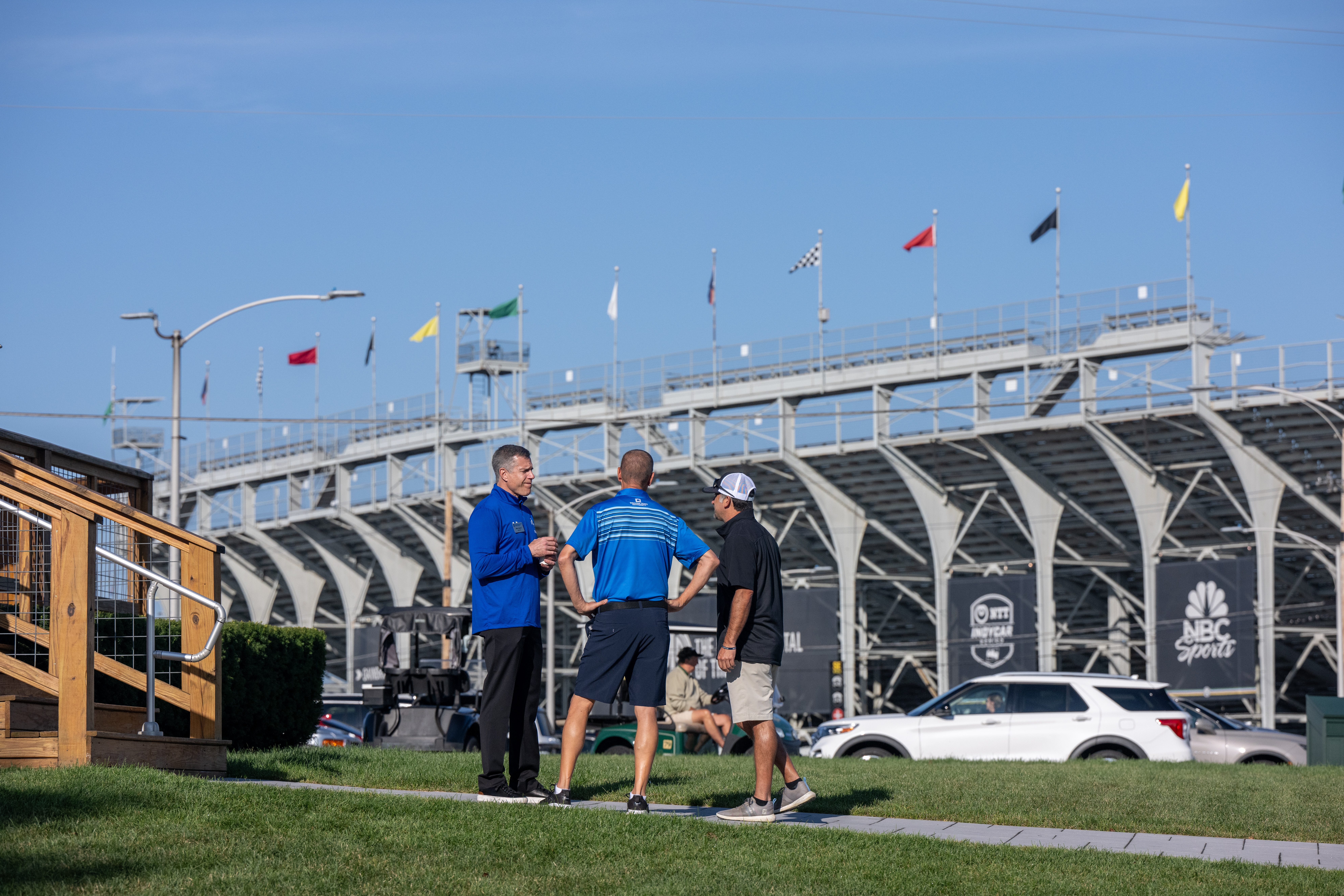 Three men in casual sportswear standing and talking on a lawn with a grandstand and flags in the background under a clear blue sky.