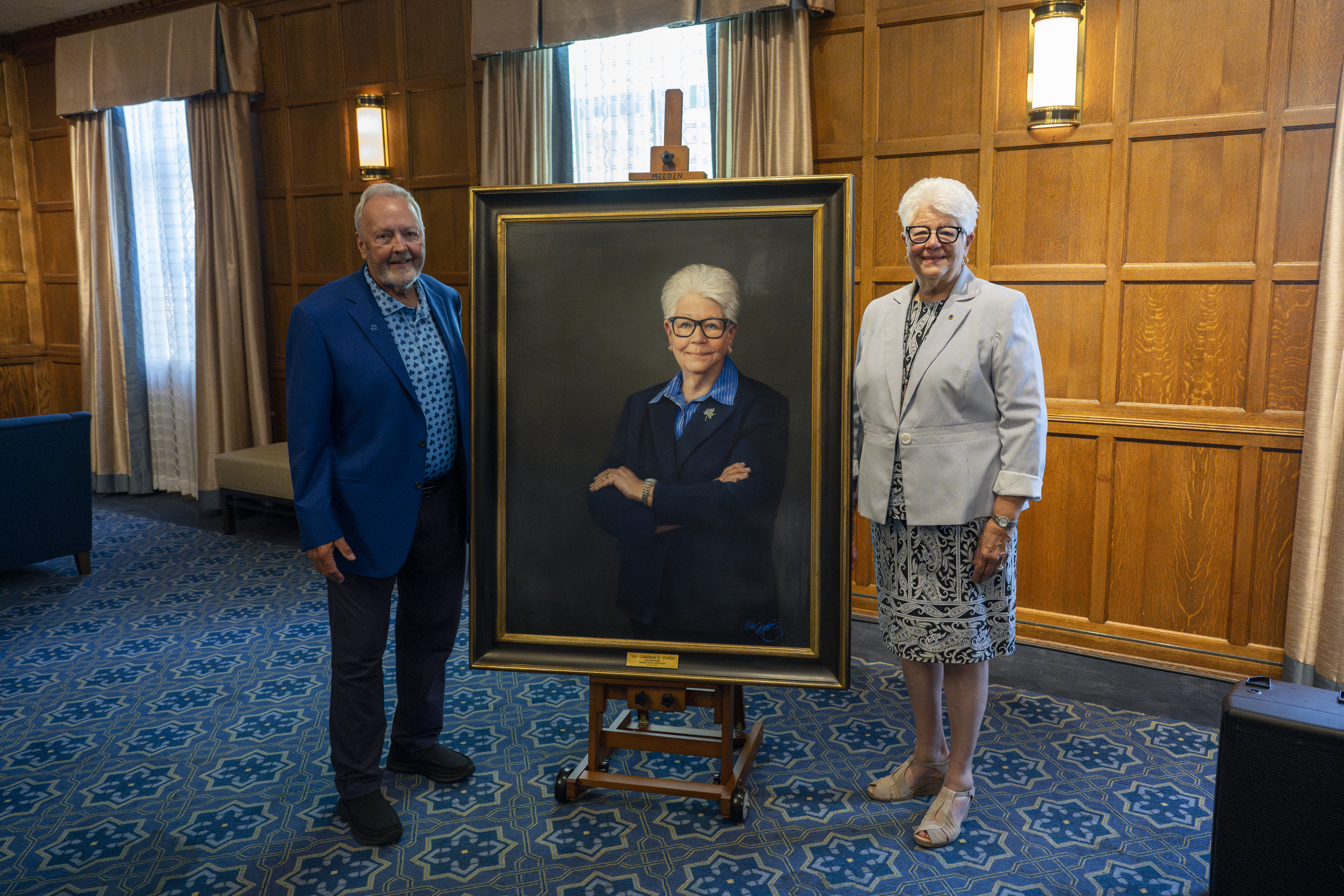 Man and woman stand on either side of a framed portrait of a woman with white hair and glasses in a wood-paneled room with blue patterned carpet.