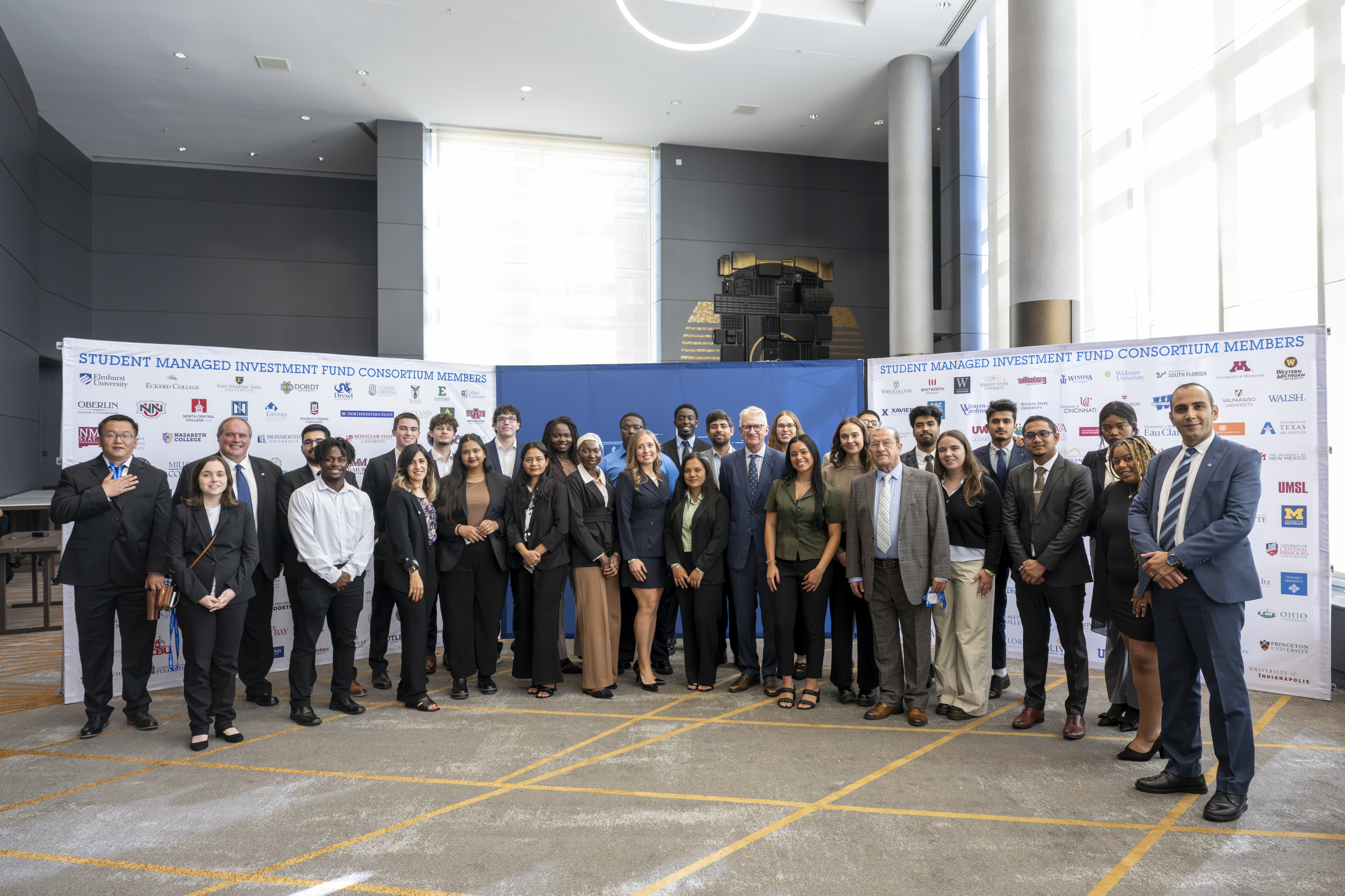 Group of diverse young adults and professionals in business attire standing in front of a conference banner with university logos indoors.