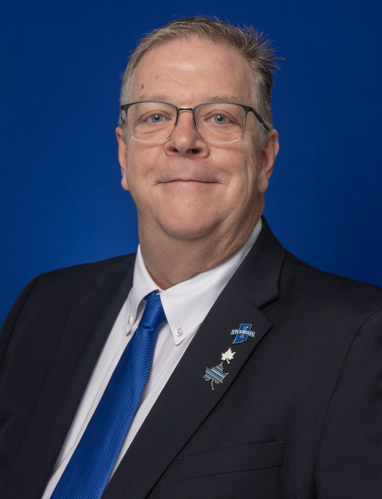 Middle-aged man wearing glasses, a dark suit, white shirt, and blue tie, posed against a solid dark blue background.