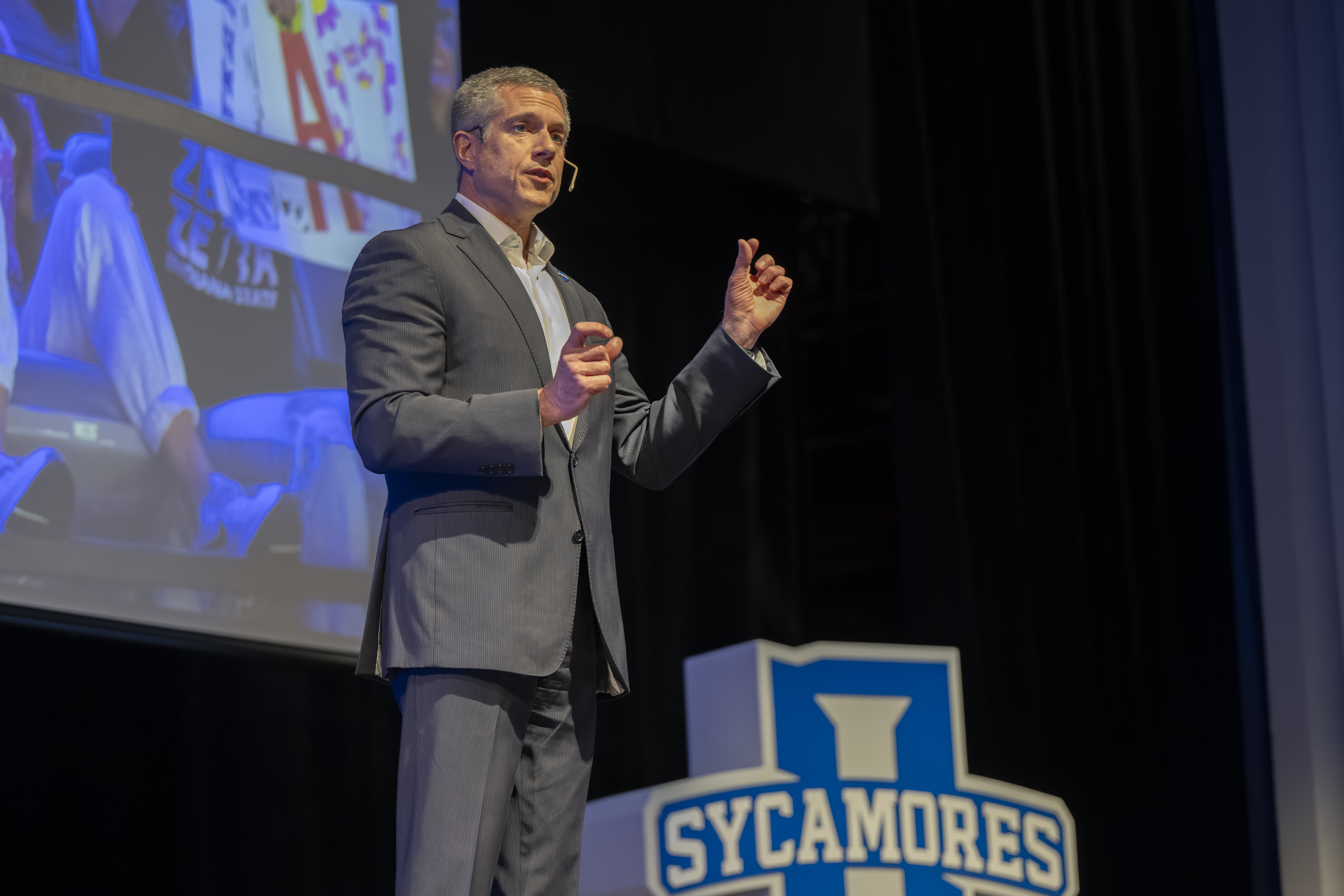 Man in gray suit speaking on stage with Sycamores logo visible near podium and a large screen behind him.