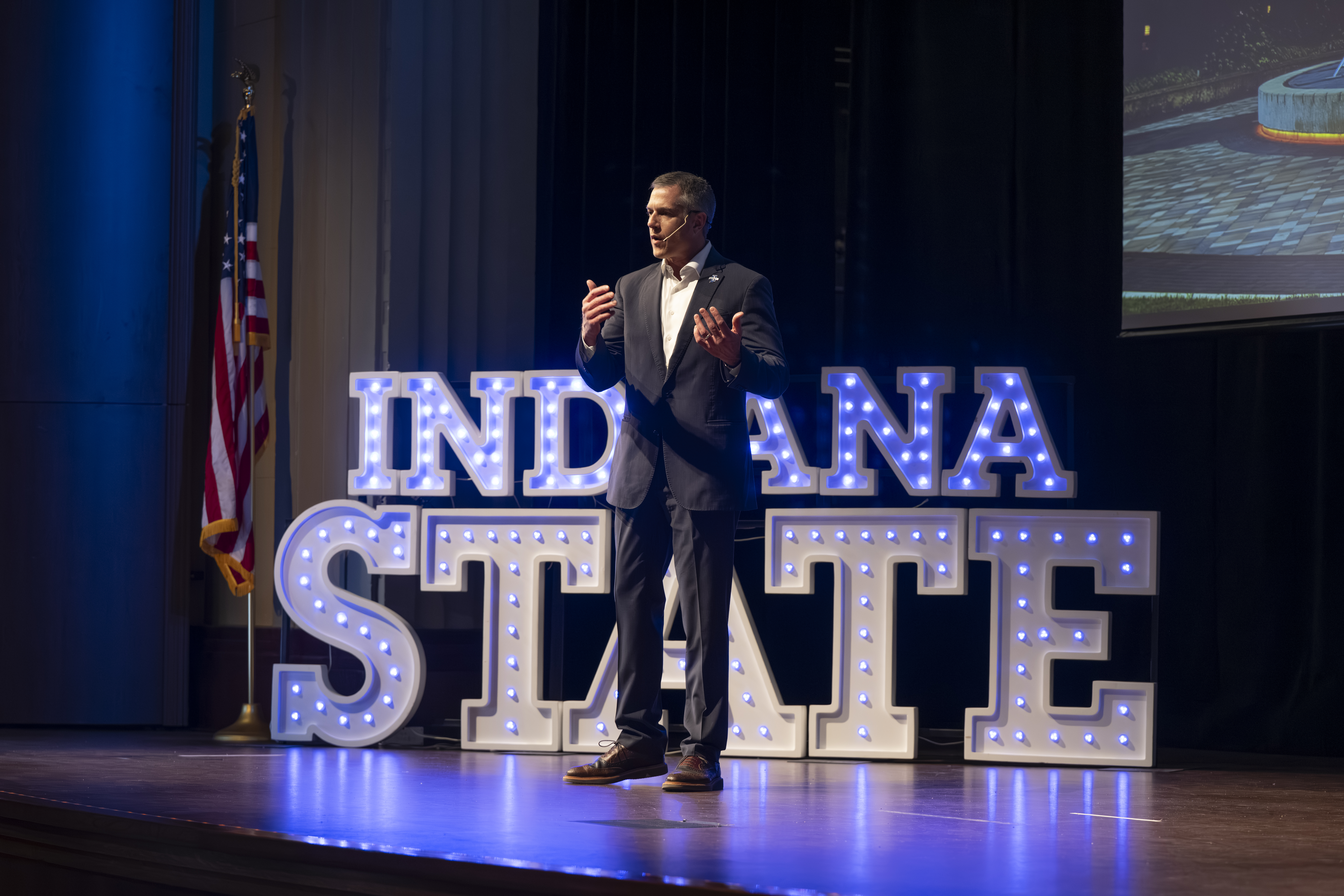 Man in suit speaking on stage with lit Indiana State sign and American flag behind him during university event.
