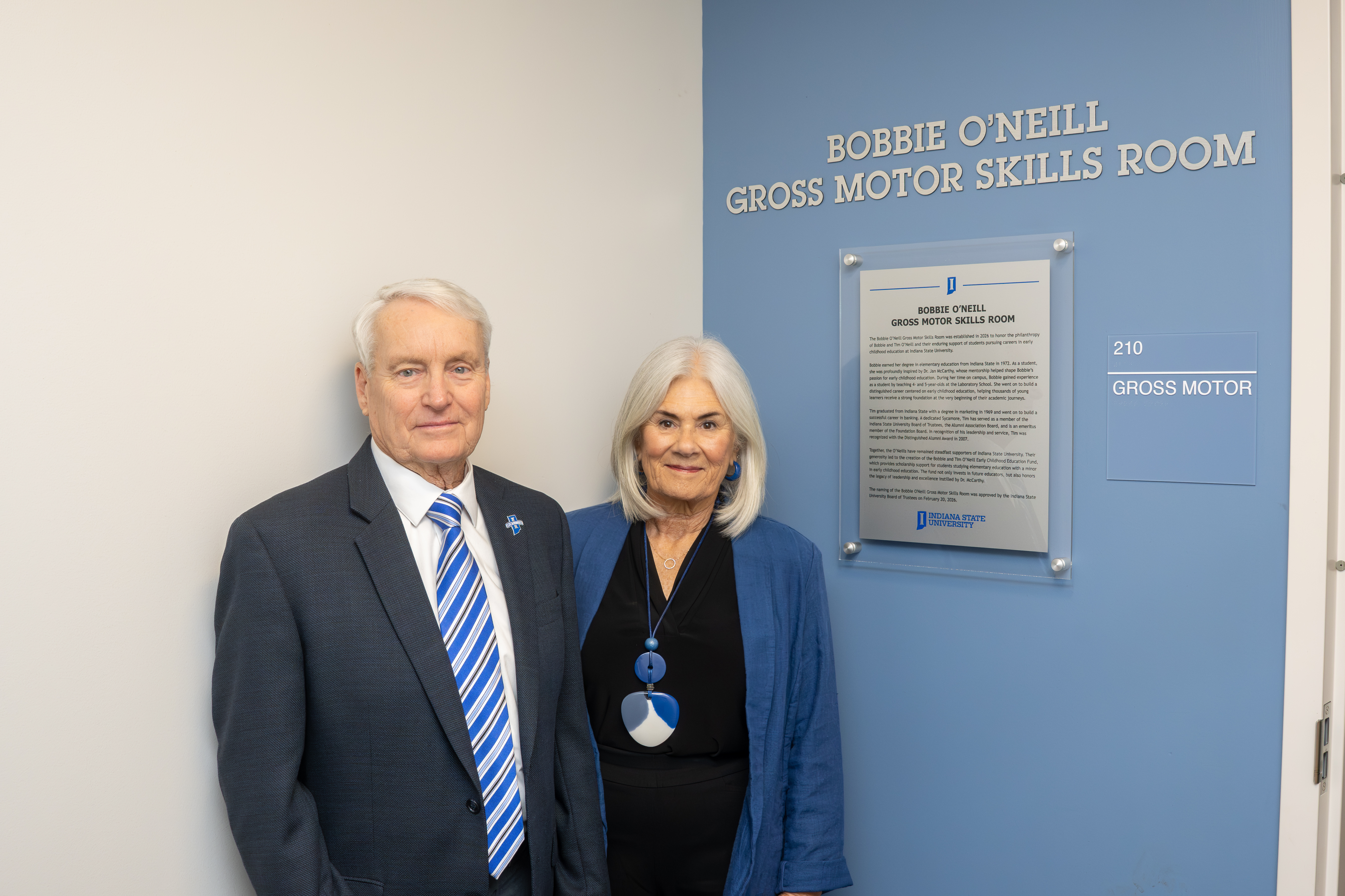 Two people stand beside a blue wall with a sign reading Bobbie O'Neill Gross Motor Skills Room at Indiana State University.