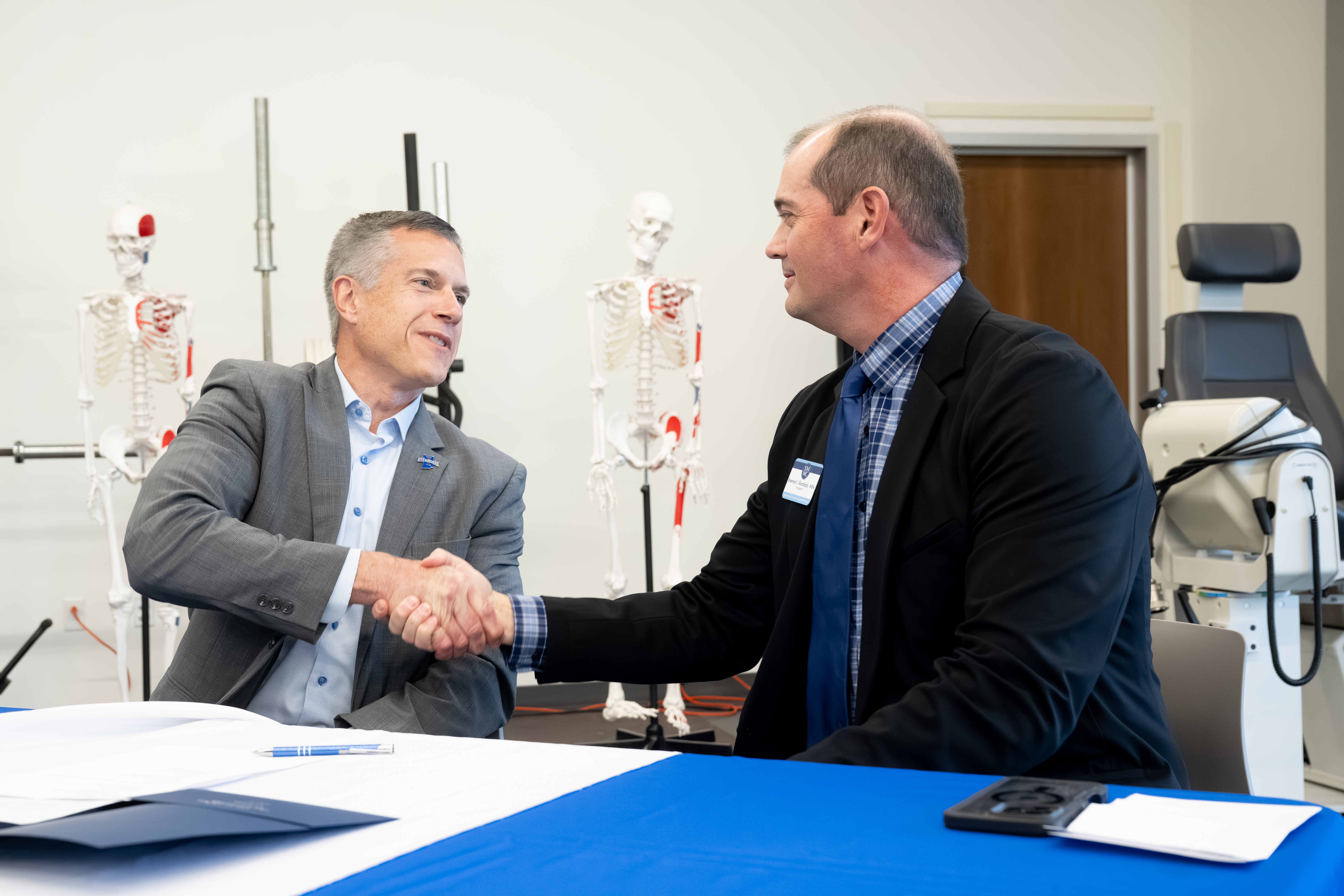 Two men in business attire shaking hands across a table with documents and anatomical skeleton models in the background.