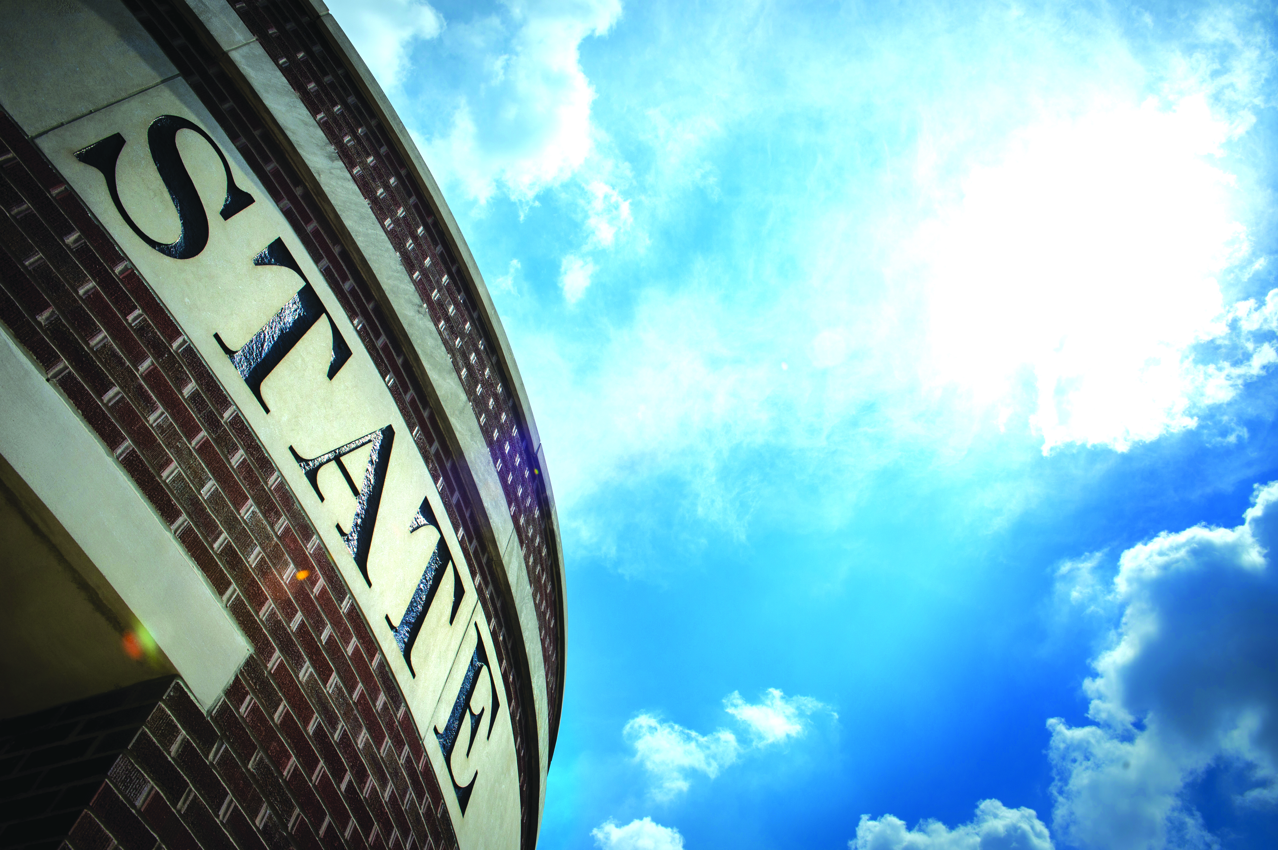 Curved brick building facade with large letters spelling STANE against a bright blue sky with clouds.