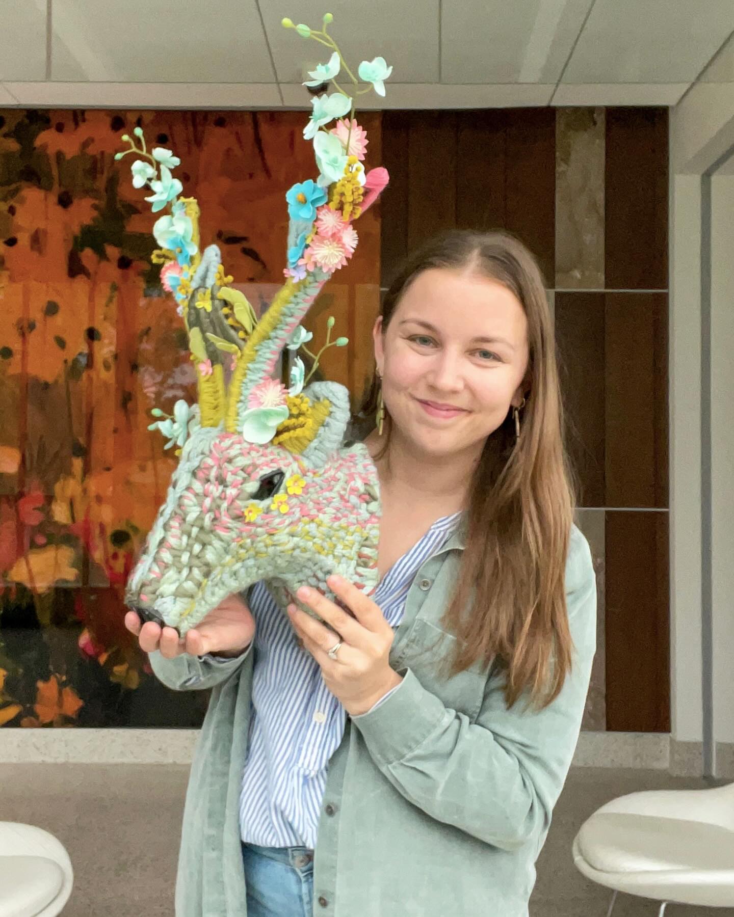 Young woman holding colorful woven deer head sculpture with floral antlers, standing indoors in front of an abstract orange and brown wall panel.