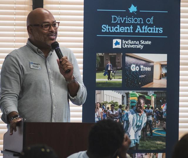 Man with glasses speaking into a microphone next to a banner for Indiana State University Division of Student Affairs with images of campus life.