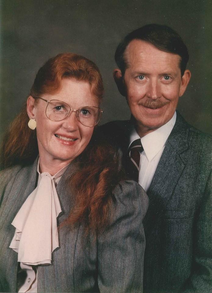 Man with mustache and brown hair in a gray suit and striped tie stands next to woman with long red hair, glasses, and gray jacket with cream blouse.