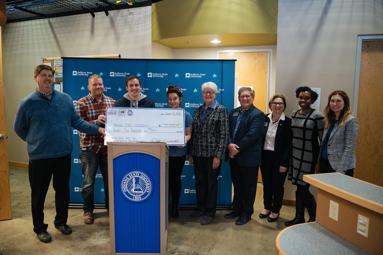 A group of nine people stand smiling behind a podium with an Indiana State University logo, holding a large ceremonial check for $25,000.