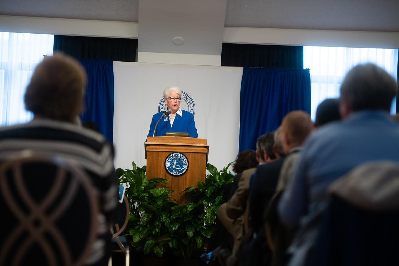 Woman in blue jacket speaking at a podium with Indiana State University seal, addressing an audience in a conference room with blue curtains and plants.