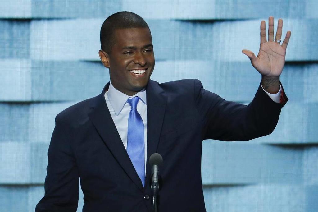 Man in dark suit and blue tie smiling and waving with right hand while standing behind a microphone against a blue-gray background.