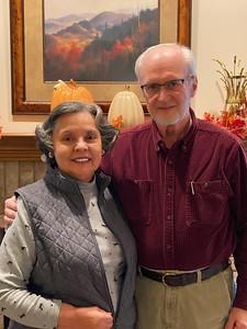 Smiling elderly couple standing indoors with autumn decor and a mountain landscape painting in the background.