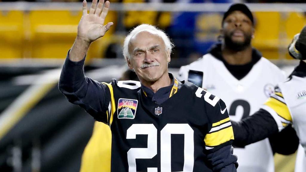 Smiling elderly man with white hair waving, wearing a black and yellow NFL jersey with the number 20 at a sports event.
