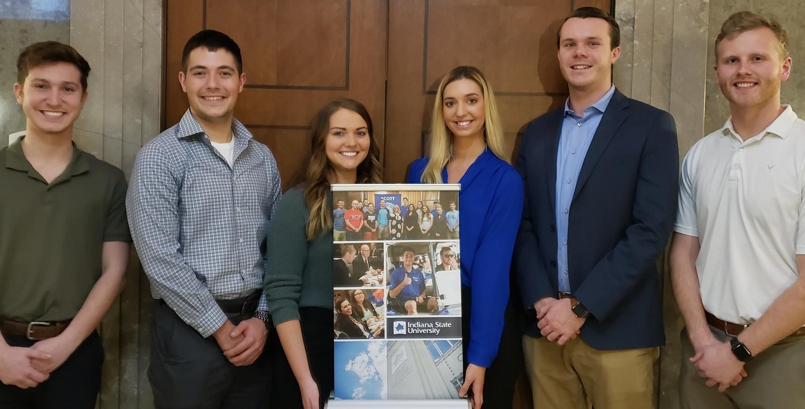 Six Indiana State University students standing indoors in front of a wood-paneled wall holding a vertical banner with Indiana State University branding.