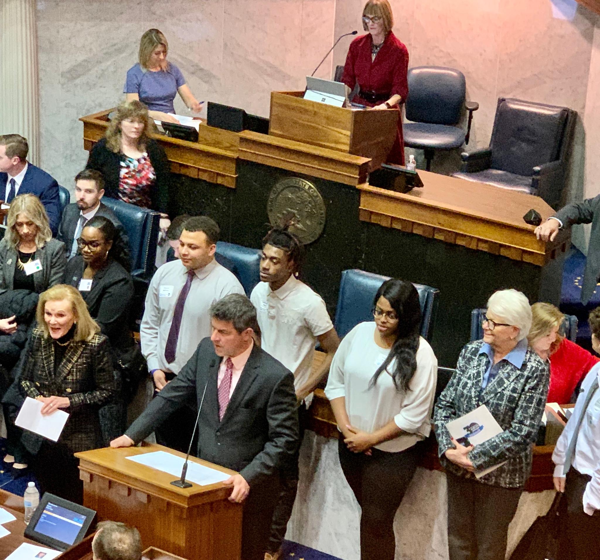 Group of people standing and seated around a podium on a marble senate floor, one man speaking at the podium, several holding papers, a woman standing behind th