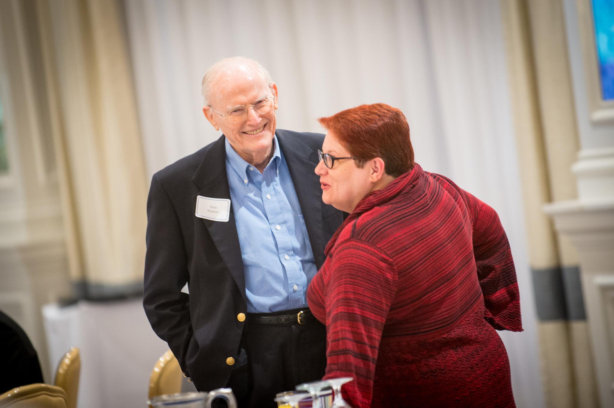Older man in black blazer and blue shirt smiling, talking with a woman in red striped sweater in an indoor event space.
