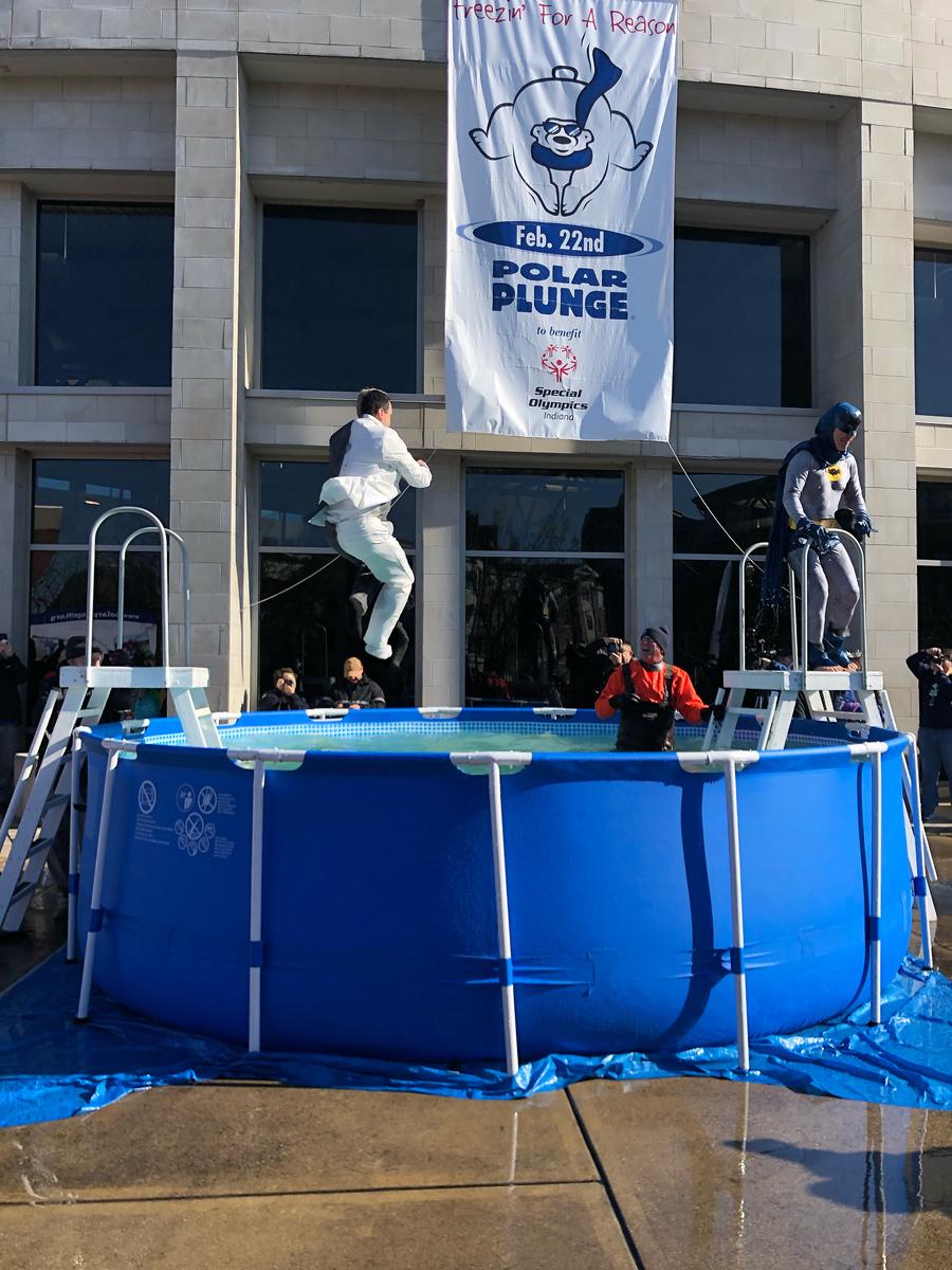 Two people in costumes jump into a blue above-ground pool outside a large building with a Polar Plunge banner for Special Olympics Indiana hanging above.