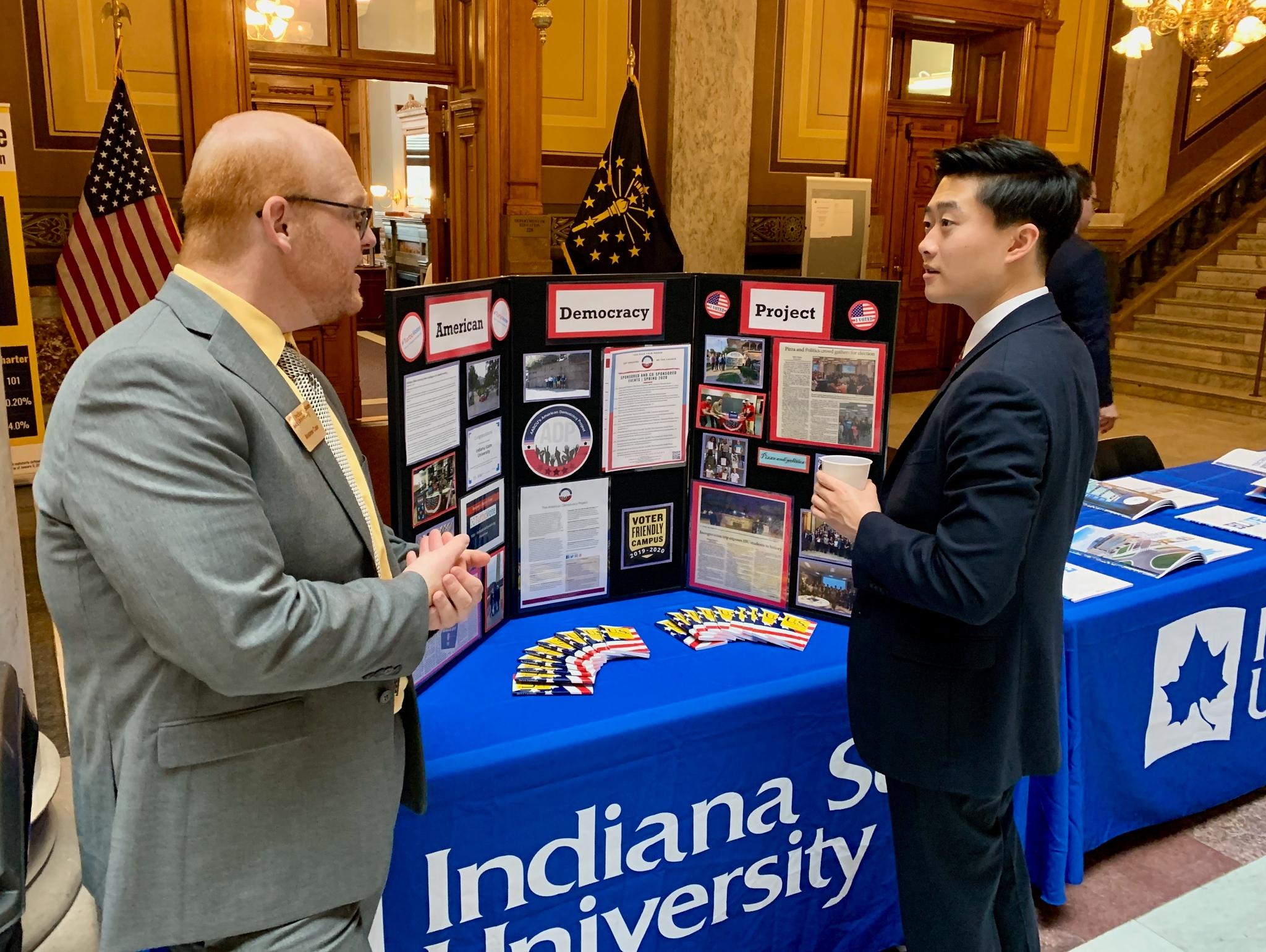Two men in suits talk beside an Indiana State University display table with an American Democracy Project tri-fold in a government building atrium.