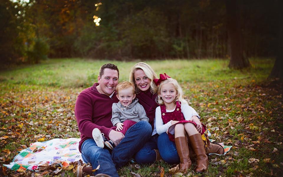 Couple and two young children sitting on a blanket in a grassy, leafy outdoor area during fall.