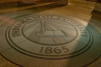 Circular Indiana State University emblem on the ground with a torch, book, and the year 1865 lit by overhead lights at night.