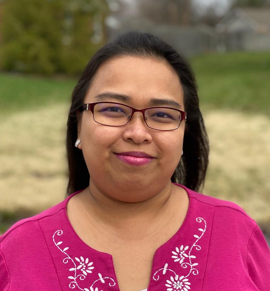 Woman wearing red glasses and a magenta top with white floral embroidery, standing outdoors with blurred grass and trees in the background.