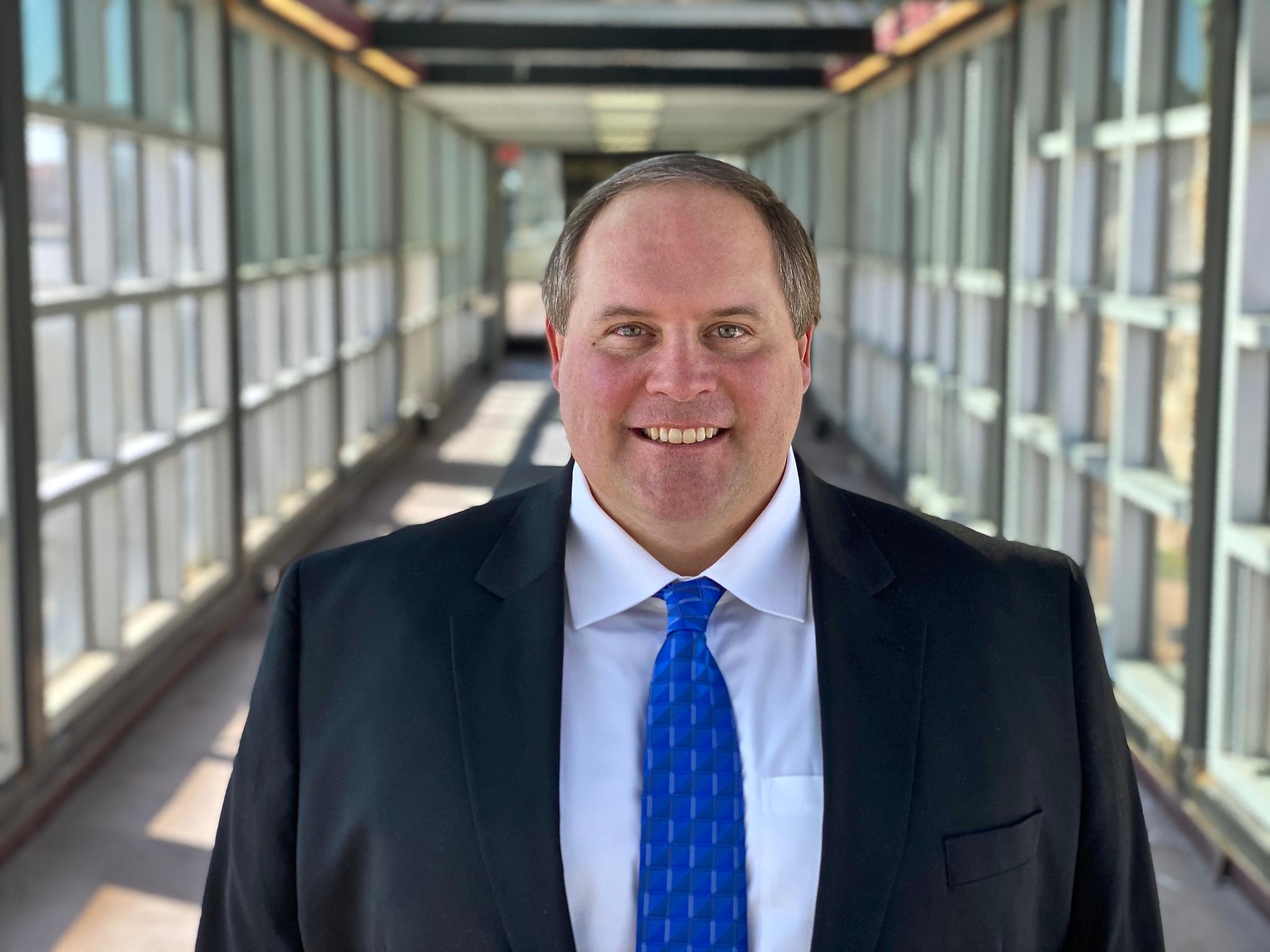 Man in business suit with blue tie smiling, standing in a glass-walled hallway with natural light.