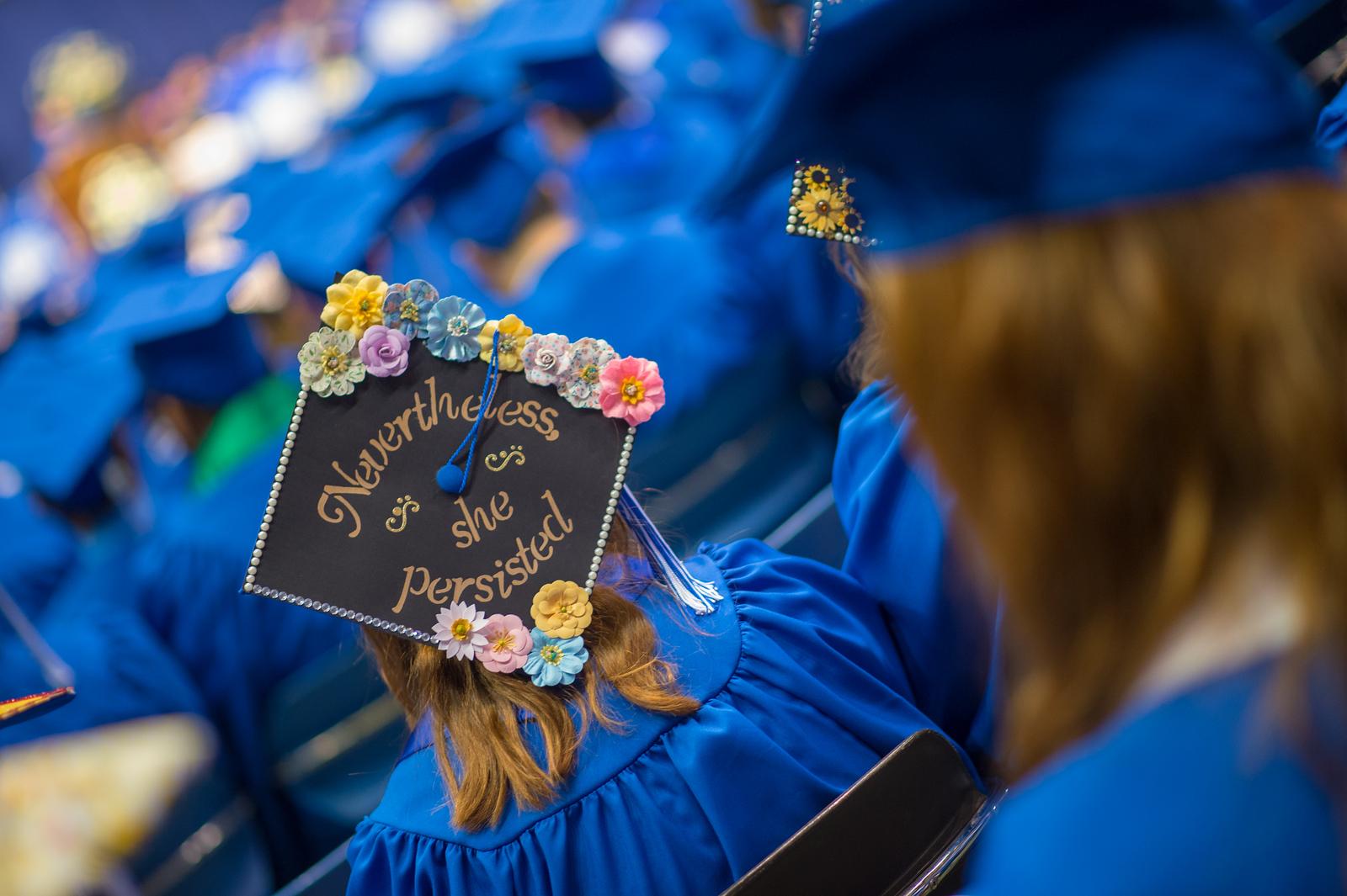 Graduates in blue caps and gowns seated, one cap decorated with flowers and the phrase nevertheless, she persisted.