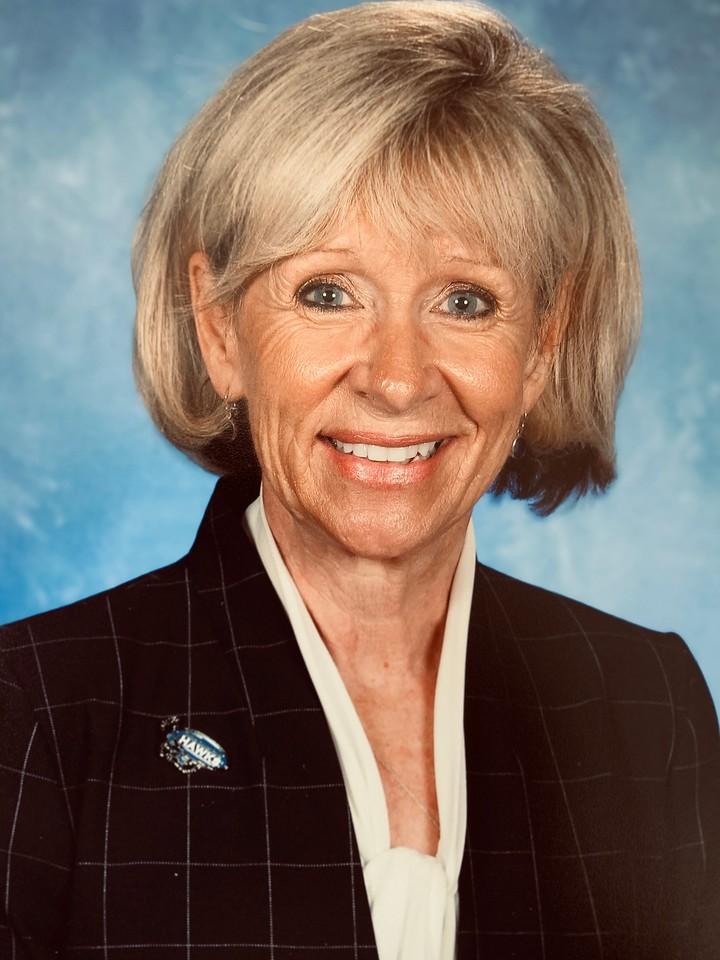 Smiling older woman with short blonde hair wearing a dark checkered blazer and light-colored blouse against a blue studio backdrop.