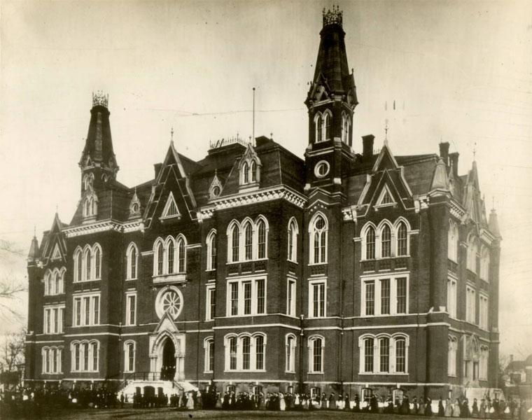 Historic Indiana State University building with tall spires and detailed Gothic architecture, surrounded by people standing along the front entrance.