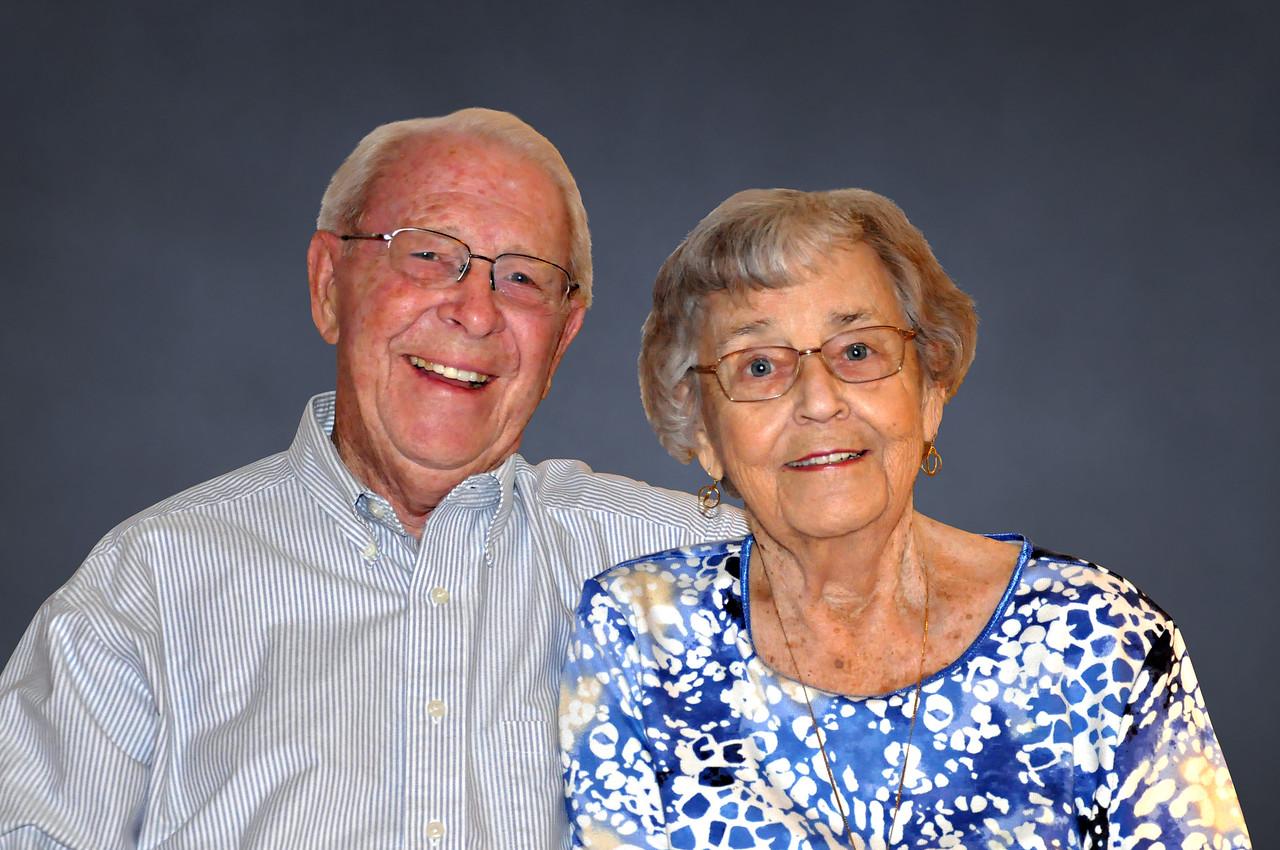 Elderly man and woman smiling, both wearing glasses, man in striped button-up shirt, woman in blue and white patterned top against plain gray background.