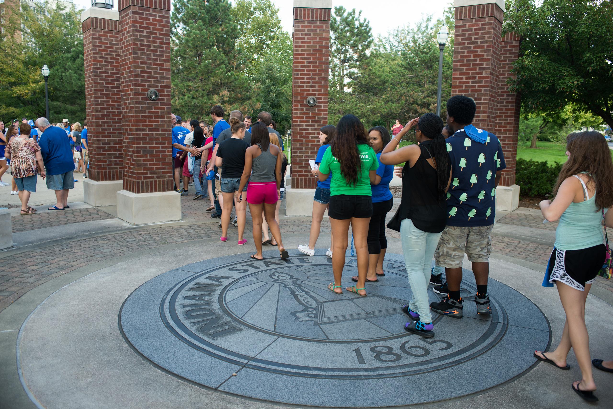 Group of young people standing in a brick-pillar archway on a circular Indiana State University emblem on the pavement.
