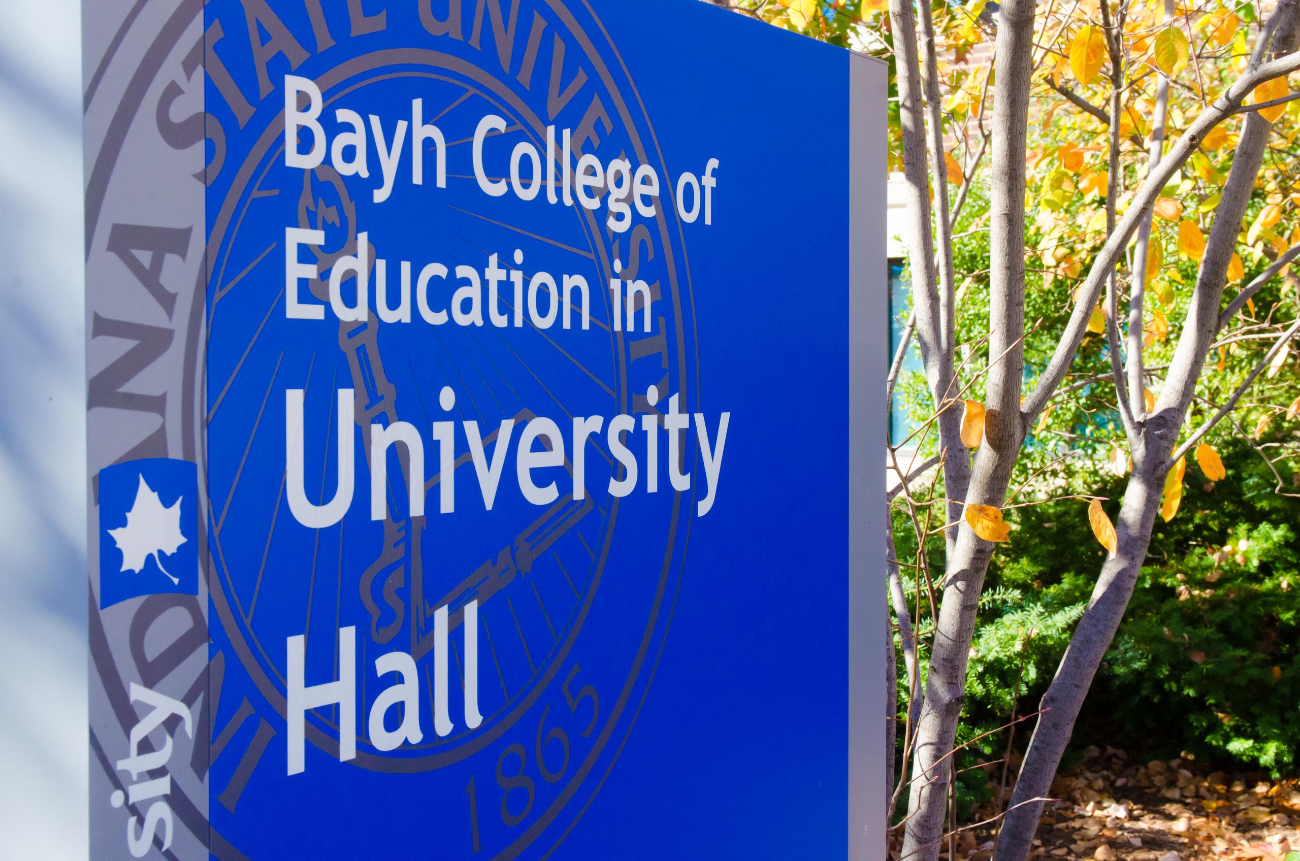 Blue sign showing Bayh College of Education at Indiana State University with part of a tree and bushes with yellow leaves nearby.