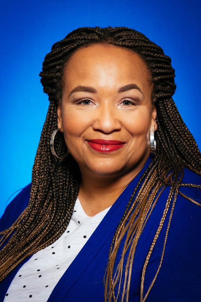 Woman with long braided hair, wearing silver hoop earrings, red lipstick, a white polka dot shirt, and a blue jacket, smiling against a blue background.